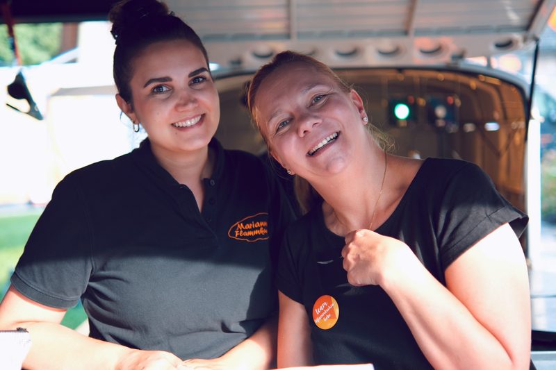 Zwei Mitarbeitende lachen beim Catering-Einsatz vor dem Ausgabewagen in die Kamera