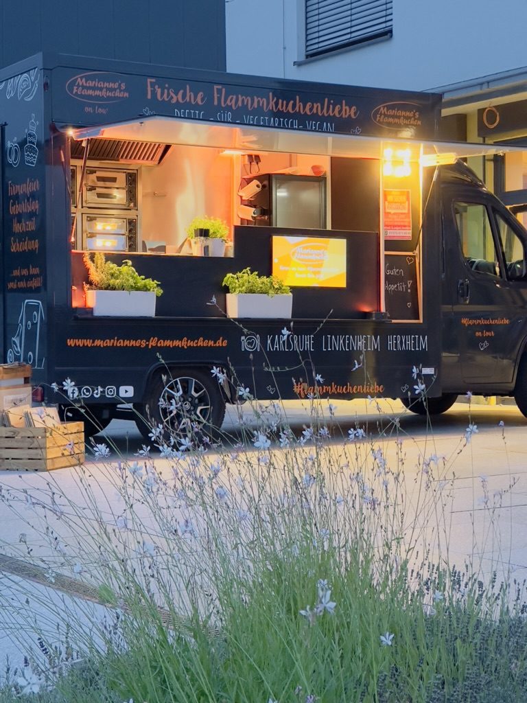 Der Foodtruck mit geöffneter Ausgabeklappe am Abend, dahinter die Flammkuchenöfen; im Vordergrund blühender Lavendel